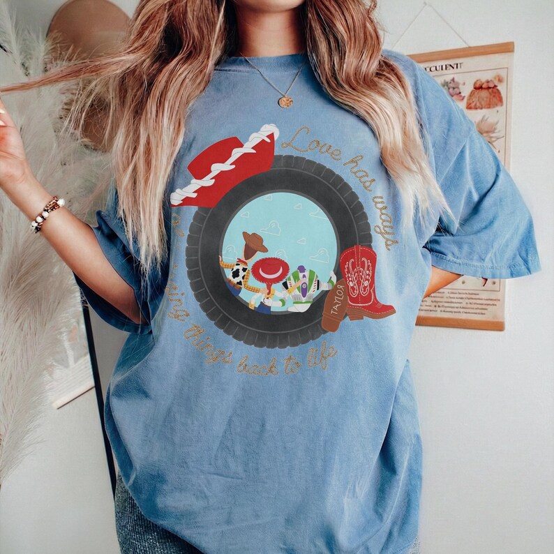 May include: A blue short-sleeve t-shirt featuring a graphic of a black tire framing cartoon characters, a red cowboy hat, and a red cowboy boot. The text "Love has ways of bringing things back to life" is printed in gold cursive around the tire graphic.
