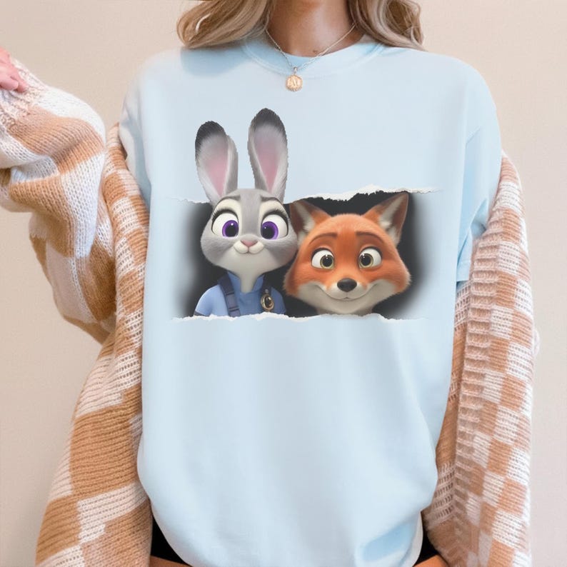 K&ouml;nnte beinhalten: Hellblaues Sweatshirt mit einem Aufdruck von Judy Hopps und Nick Wilde aus Zoomania. Die Charaktere sind durch einen zerrissenen Papiereffekt dargestellt. Das Sweatshirt besteht aus weichem Material und ist ideal f&uuml;r den Alltag.