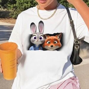 K&ouml;nnte beinhalten: Wei&szlig;es T-Shirt mit einer Grafik von Judy Hopps und Nick Wilde aus Zoomania. Die Zeichentrickfiguren sind in einem zerrissenen Papiereffekt dargestellt. Das Shirt besteht aus einem weichen Material.