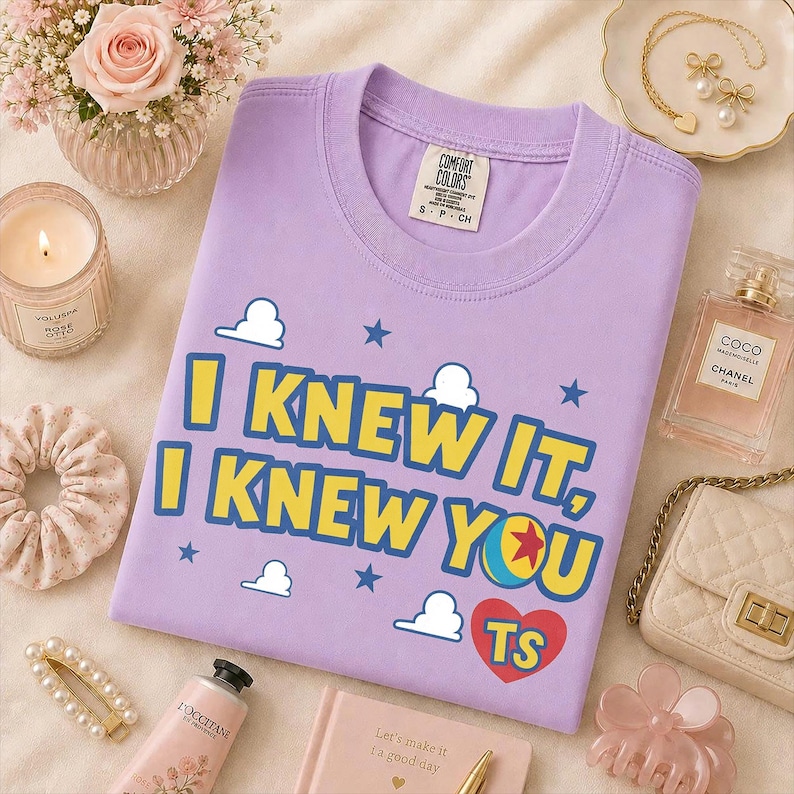 Pu&ograve; includere: Una t-shirt a maniche corte color viola chiaro con il testo giallo I KNEW IT, I KNEW YOU, un cuore rosso contenente le lettere TS, stelle blu e grafiche di nuvole bianche. La maglietta presenta un'etichetta del marchio Comfort Colors.