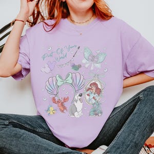 May include: A light purple t-shirt featuring a graphic print with the text Part Of Your World, a pair of seashell ears, a bow, bubbles, a crab, a fish, a dog, and a framed illustration of a red-haired character in a blue dress.