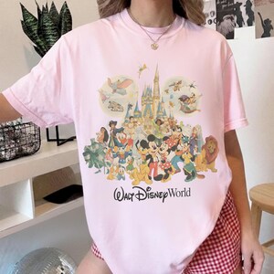 May include: A light pink t-shirt featuring a colorful graphic of Disney characters and a castle. The words "Walt Disney World" are printed below the graphic. The shirt has short sleeves and a crew neck.