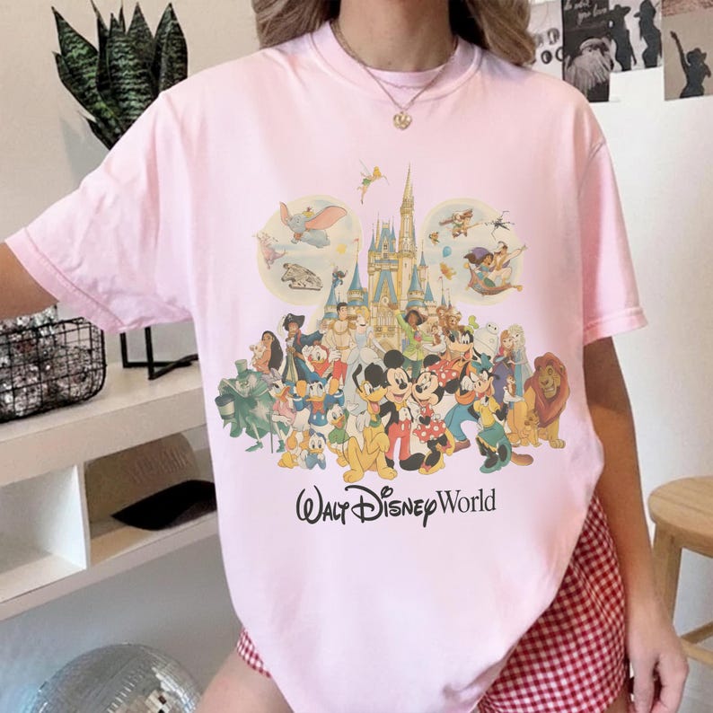 May include: A light pink t-shirt featuring a colorful graphic of Disney characters and a castle. The words "Walt Disney World" are printed below the graphic. The shirt has short sleeves and a crew neck.