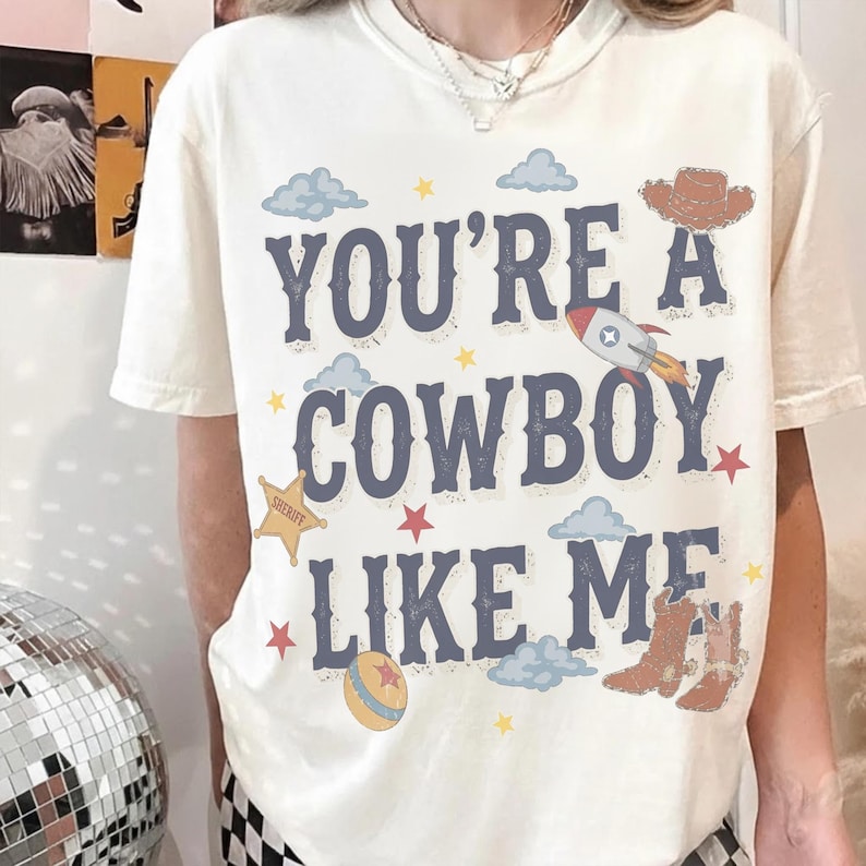 May include: A cream-colored short-sleeve t-shirt featuring the blue text "YOU'RE A COWBOY LIKE ME" surrounded by graphic illustrations of a brown cowboy hat, a rocket ship, a yellow sheriff star, a ball, clouds, and small red and yellow stars.