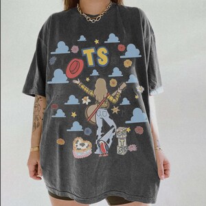 May include: A charcoal grey short-sleeve t-shirt featuring a graphic print of a person with a guitar, a red hat, blue clouds, flowers, and stars. The text TS is printed in yellow and blue, with additional blocks displaying the letters T and S.