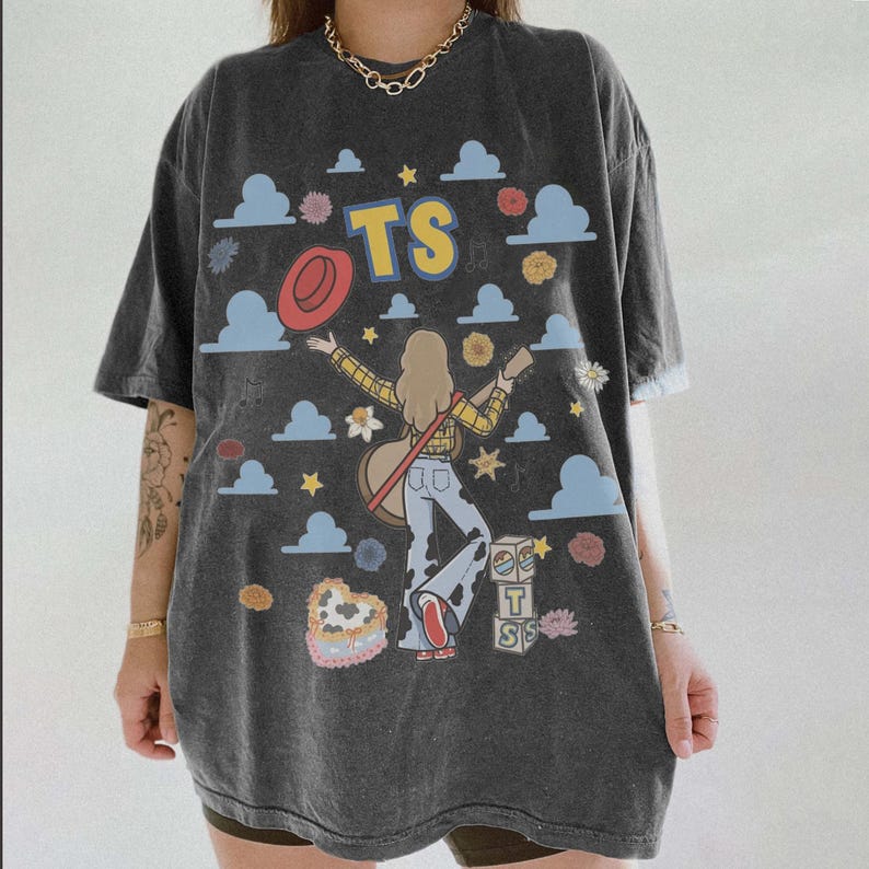 May include: A charcoal grey short-sleeve t-shirt featuring a graphic print of a person with a guitar, a red hat, blue clouds, flowers, and stars. The text TS is printed in yellow and blue, with additional blocks displaying the letters T and S.