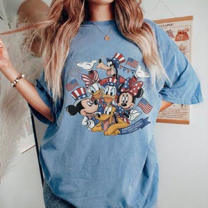 May include: Pale blue t-shirt with a graphic of Disney characters Mickey Mouse, Minnie Mouse, Donald Duck, Daisy Duck, Goofy, and Pluto, celebrating the USA's 250th anniversary. The design includes American flags and fireworks. The text on the shirt reads "America 250th USA Anniversary 1776-2026".