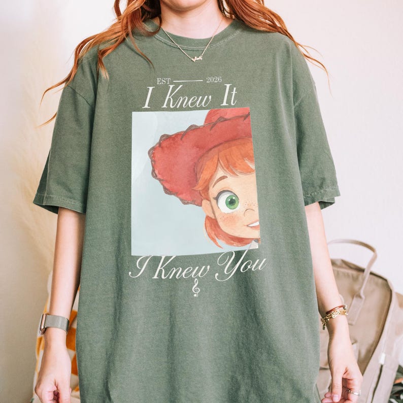 May include: An olive green short-sleeve t-shirt featuring a graphic print of a cartoon character with red hair and a red hat. The text above the graphic reads EST 2026 I Knew It and the text below the graphic reads I Knew You with a musical note symbol.