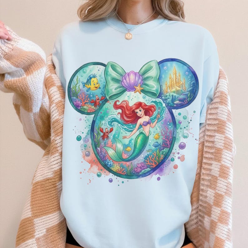 May include: Light blue sweatshirt featuring a colorful illustration of Ariel from The Little Mermaid. The design incorporates a Mickey Mouse ear shape with underwater scenes, a bow, and a castle. The artwork includes Flounder, Sebastian, and other sea creatures.