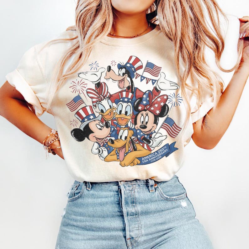 May include: Cream-coloured t-shirt with a graphic of Disney characters Mickey Mouse, Minnie Mouse, Donald Duck, Daisy Duck, Goofy, and Pluto, in patriotic outfits. The design includes American flags and fireworks, with text reading "America 250th USA Anniversary 1776-2026".