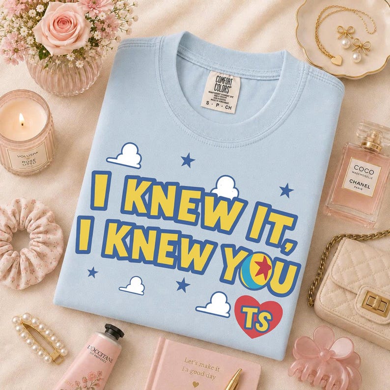 Pu&ograve; includere: Una t-shirt azzurra con il testo giallo "I KNEW IT, I KNEW YOU" con contorni blu, accompagnata da grafiche di nuvole bianche, stelle blu, un'icona a forma di palla colorata e un cuore rosso contenente le lettere "TS" stampate sul davanti.