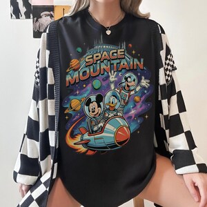 Może przedstawiać: Czarny t-shirt z grafiką Space Mountain z Myszką Miki, Kaczorem Donaldem i Goofym w skafandrach kosmicznych jadących na rakiecie. Koszulka ma kolorowe tło sceny kosmicznej. Na koszulce noszony jest czarno-biały kardigan w kratę.
