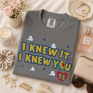 Pu&ograve; includere: Una t-shirt grigio antracite a girocollo con il testo giallo I KNEW IT, I KNEW YOU, una grafica a palla colorata, un cuore rosso con le lettere TS, piccole nuvole bianche e stelle blu stampate sul davanti, con un'etichetta del marchio Comfort Colors.