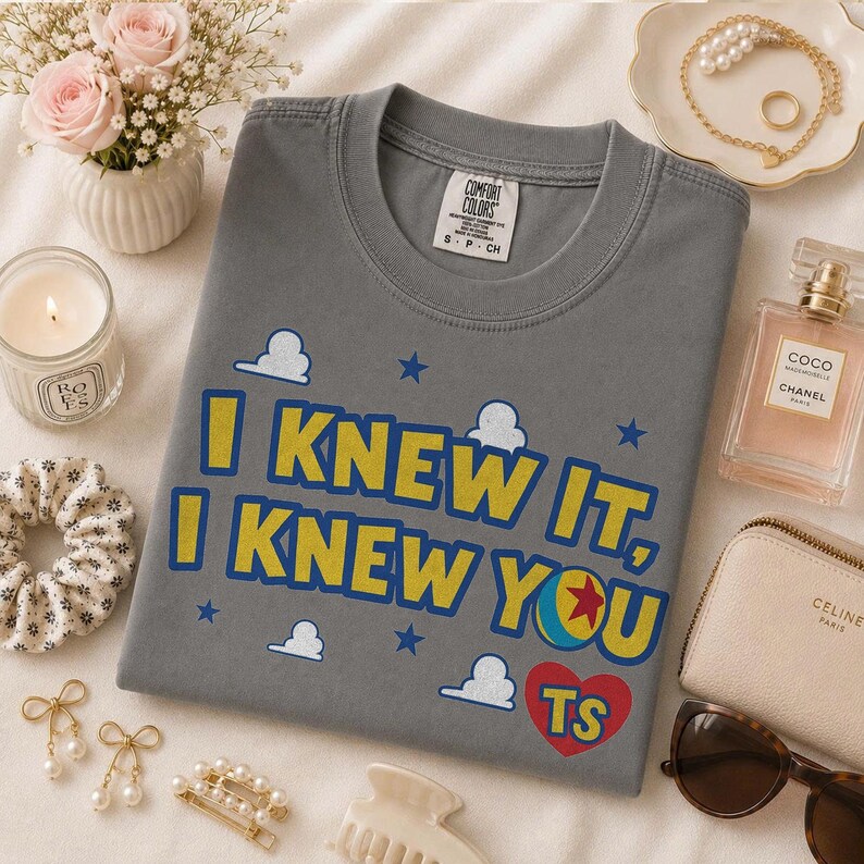 Pu&ograve; includere: Una t-shirt grigio antracite a girocollo con il testo giallo I KNEW IT, I KNEW YOU, una grafica a palla colorata, un cuore rosso con le lettere TS, piccole nuvole bianche e stelle blu stampate sul davanti, con un'etichetta del marchio Comfort Colors.
