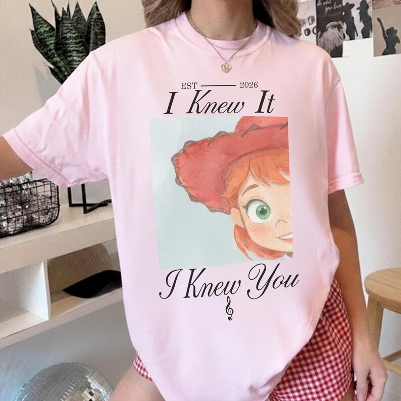 May include: A light pink t-shirt featuring a graphic of a cartoon character with red hair and a red hat. The text reads EST 2026 I Knew It I Knew You with a treble clef symbol below the text. The character has green eyes and freckles.