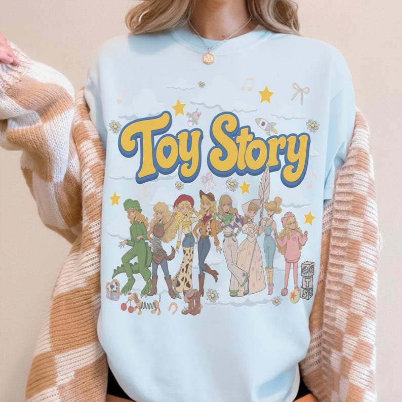 May include: A light blue crewneck sweatshirt featuring the yellow text Toy Story above a graphic illustration of several characters dressed in various outfits, including a dinosaur costume, cowgirl attire, and space-themed clothing, with stars.