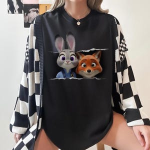 K&ouml;nnte beinhalten: Schwarzes Sweatshirt mit Rundhalsausschnitt und einer Grafik von Judy Hopps und Nick Wilde aus Zootopia. Das Sweatshirt wird mit einer schwarz-wei&szlig; karierten Strickjacke kombiniert. Die Charaktere erscheinen aus einer zerrissenen Tasche.