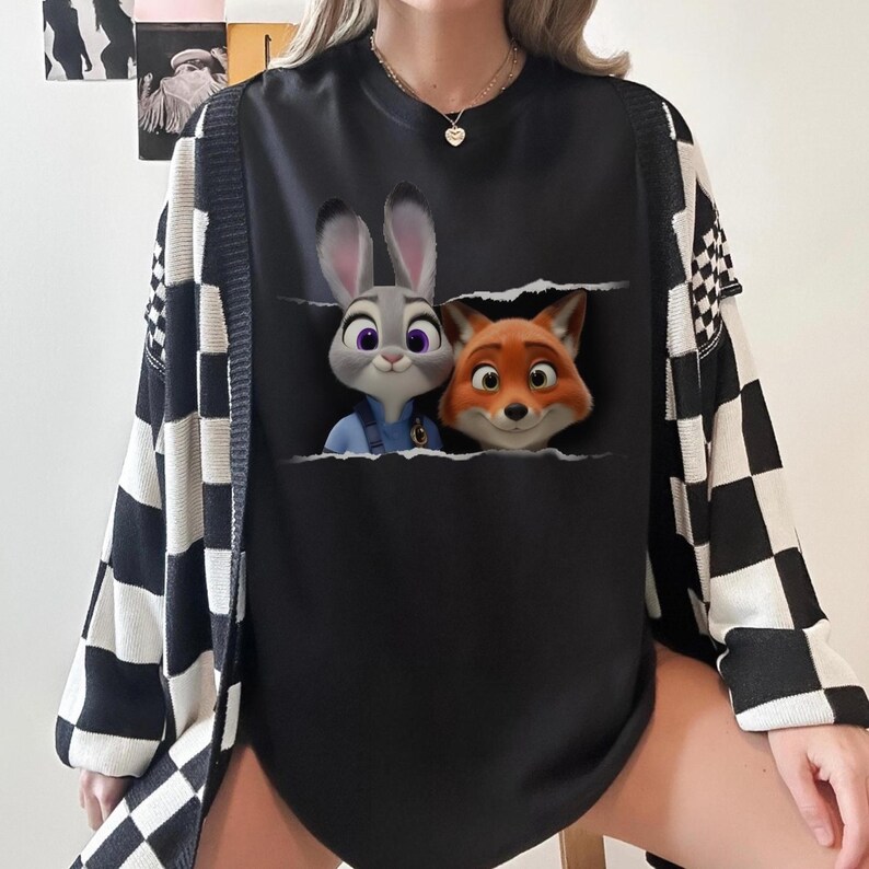 K&ouml;nnte beinhalten: Schwarzes Sweatshirt mit Rundhalsausschnitt und einer Grafik von Judy Hopps und Nick Wilde aus Zootopia. Das Sweatshirt wird mit einer schwarz-wei&szlig; karierten Strickjacke kombiniert. Die Charaktere erscheinen aus einer zerrissenen Tasche.