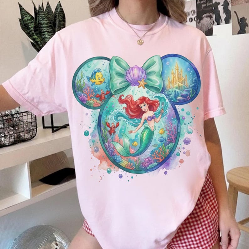 May include: Light pink t-shirt featuring a colorful graphic of Ariel the mermaid inside a Mickey Mouse head design. The design includes a teal bow, a seashell, and images of Flounder, a castle, and other sea creatures. The shirt is made of a soft material.