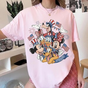 May include: A pale pink t-shirt with a graphic of Disney characters Mickey Mouse, Minnie Mouse, Donald Duck, Daisy Duck, Goofy, and Pluto, in patriotic outfits. The design includes American flags and fireworks, celebrating the USA's 250th anniversary.