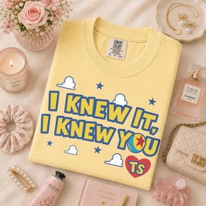 Pu&ograve; includere: Una t-shirt a girocollo giallo pallido con il testo blu I KNEW IT, I KNEW YOU, un cuore rosso contenente le lettere TS, circondato da nuvole bianche e stelle blu. La maglietta presenta un'etichetta del marchio Comfort Colors sul colletto.