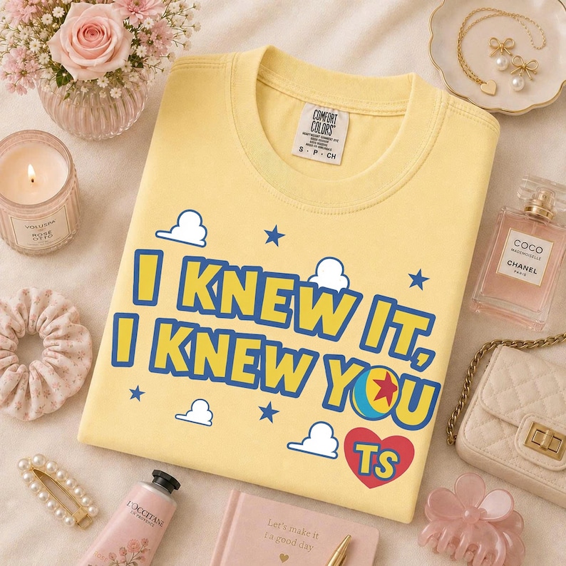 Pu&ograve; includere: Una t-shirt a girocollo giallo pallido con il testo blu I KNEW IT, I KNEW YOU, un cuore rosso contenente le lettere TS, circondato da nuvole bianche e stelle blu. La maglietta presenta un'etichetta del marchio Comfort Colors sul colletto.