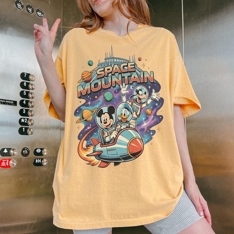 Może przedstawiać: Ż&oacute;łta koszulka z grafiką Space Mountain z Myszką Miki, Kaczorem Donaldem i Goofym w skafandrach kosmicznych na rakiecie. Koszulka ma retro design z planetami i gwiazdami.