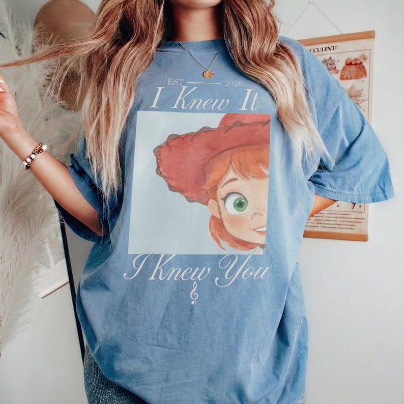 May include: A blue short-sleeve t-shirt featuring a graphic of a character with red hair and a red hat. The text reads EST 2026, I Knew It, and I Knew You. The design is centered on the chest of the shirt, which has a relaxed, loose-fitting silhouette.