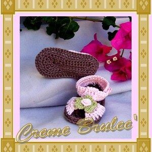 May include: A pair of crocheted baby shoes. One shoe is brown and the other is pink with a green flower embellishment. The shoes are on a white fabric background.