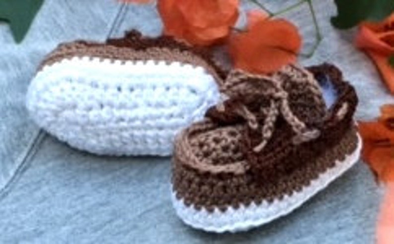 Puede incluir: Un par de botitas de beb&eacute; de ganchillo de color marr&oacute;n y blanco con cordones. Las botitas tienen una suela blanca y una parte superior marr&oacute;n con un patr&oacute;n de rayas marrones y blancas.