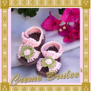 May include: A pair of pink and brown crocheted baby sandals with flower embellishments. The sandals are on a white fabric background with pink flowers.