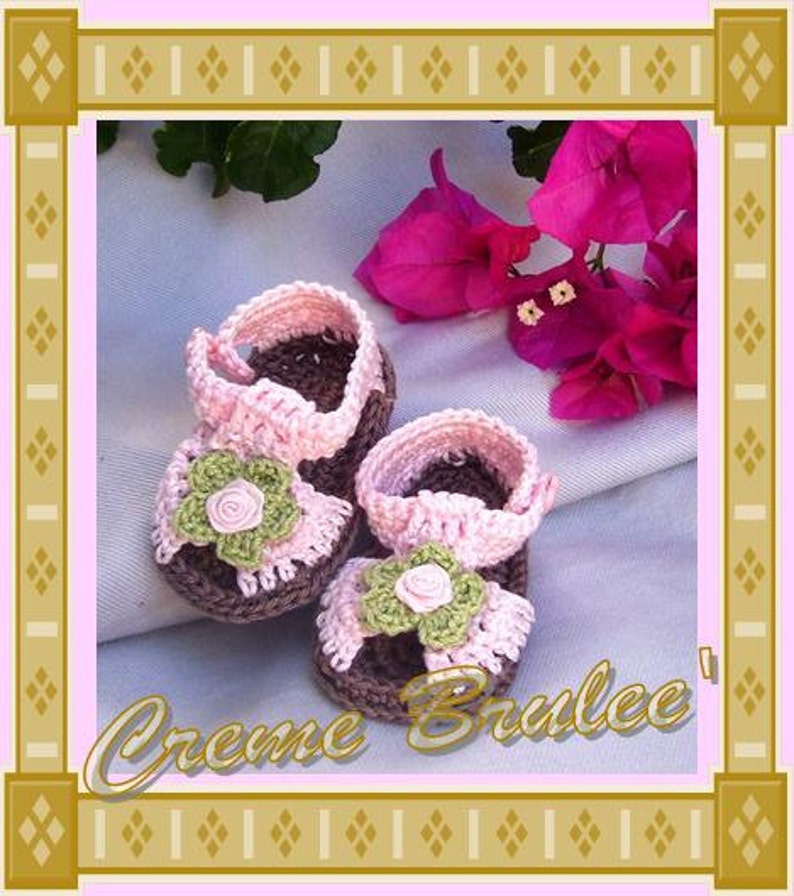 May include: A pair of pink and brown crocheted baby sandals with flower embellishments. The sandals are on a white fabric background with pink flowers.
