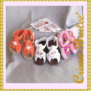 May include: Five pairs of crocheted baby sandals in orange, white, and pink with flower embellishments. The sandals are handmade and have a delicate, feminine design.