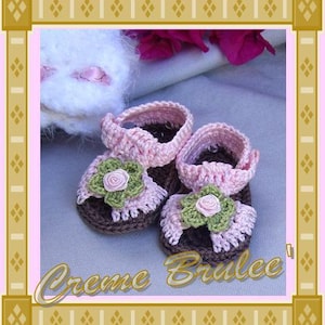 May include: A pair of pink and brown crocheted baby sandals with flower embellishments. The sandals are on a light pink fabric background.