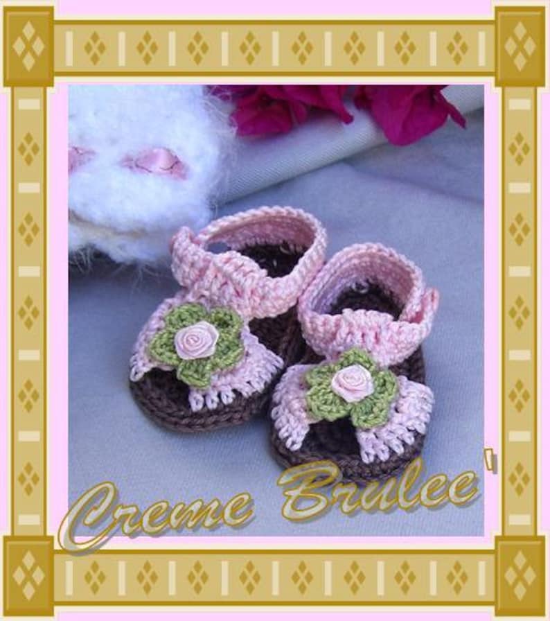 May include: A pair of pink and brown crocheted baby sandals with flower embellishments. The sandals are on a light pink fabric background.