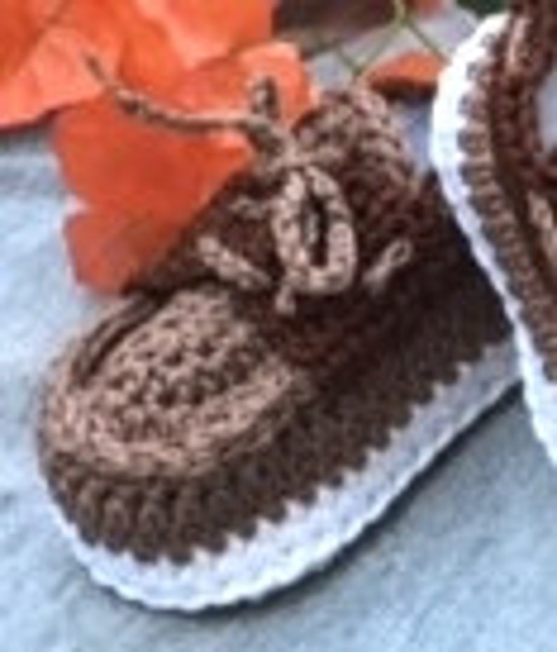 Puede incluir: Botitas de beb&eacute; de crochet marr&oacute;n y blanco con cordones. Las botitas tienen un dise&ntilde;o texturizado y un ribete blanco.