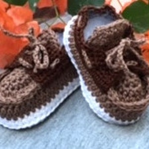 Puede incluir: Un par de zapatos de beb&eacute; de ganchillo marr&oacute;n y blanco con cordones. Los zapatos tienen una suela blanca y una parte superior marr&oacute;n.
