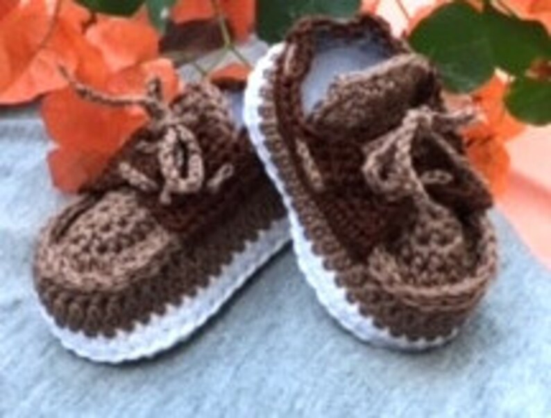 Puede incluir: Un par de zapatos de beb&eacute; de ganchillo marr&oacute;n y blanco con cordones. Los zapatos tienen una suela blanca y una parte superior marr&oacute;n.