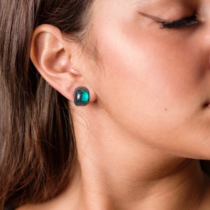 May include: A woman wearing a pair of dark green square-shaped earrings. The earrings have a clear, glassy appearance.
