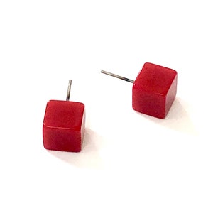 May include: A pair of red cube stud earrings. Each earring features a small, square, solid red cube with a silver-colored post. The earrings are set against a white background, highlighting their simple, geometric design.