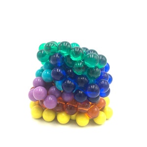 May include: A rainbow stack of colorful glass marbles. The marbles are arranged in a pyramid shape, with the largest marbles at the bottom and the smallest marbles at the top. The colors of the marbles are yellow, orange, purple, blue, and green.
