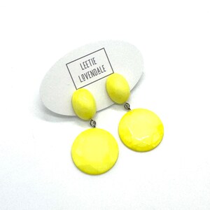 May include: Bright yellow drop earrings featuring a small oval stud attached to a larger circular pendant. The earrings are displayed on a white oval card with the text LEETIE LOVENDALE printed in black inside a rectangular border.