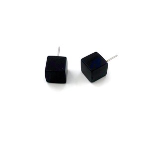 Puede incluir: Dos pendientes de botón cuadrados negros con postes plateados. Estos pendientes minimalistas tienen un diseño geométrico simple. Se presentan sobre un fondo blanco.