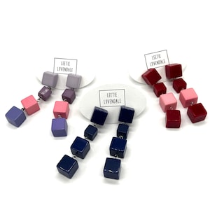 May include: Three pairs of drop earrings featuring stacked cube beads in purple, pink, navy blue, and red colourways. Each pair is attached to a white oval display card printed with the text Leetie Lovendale. The cubes have a smooth, glossy finish.