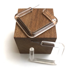 May include: A pair of clear acrylic square hoop earrings. The earrings are shown on a wooden block.