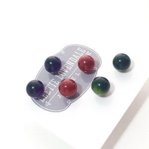 May include: Five small round glass beads in various colours, including green, red, and purple. The beads are arranged on a white surface with a grey label that reads "Lettie Lovendale Jewelry".