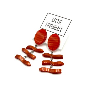 May include: A pair of orange earrings with an oval top and three curved, ribbon-like dangles. The earrings are connected with silver-colored metal. A white card with the text "LEETIE LOVENDALE" is in the background.
