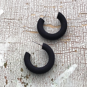 Könnte beinhalten: Ein Paar schwarze Creolen. Die Ohrringe sind aus einem dicken, matten Material gefertigt und haben ein schlichtes, modernes Design.