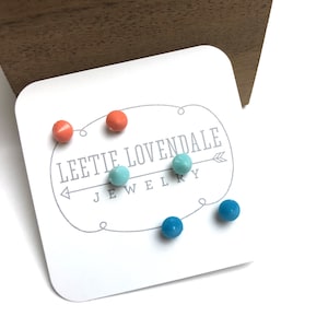 Pode incluir: Um conjunto de cinco brincos de pressão coloridos em um cartão branco com o texto "Leetie Lovendale Jewelry". Os brincos são em tons de azul e laranja.