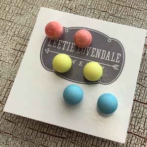 以下が含まれることがあります： 白いカードに「Leetie Luvendale Jewelry」の文字が書かれた、6ペアのカラフルなスタッドピアス。ピアスはピンク、イエロー、ブルーの色合いです。